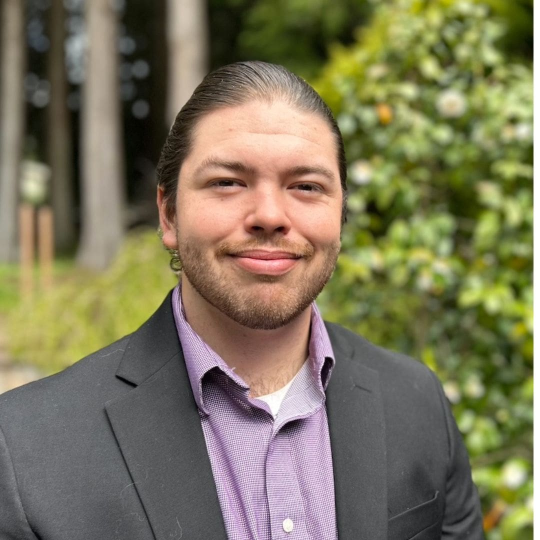 Man in suit smiling outdoors near trees.
